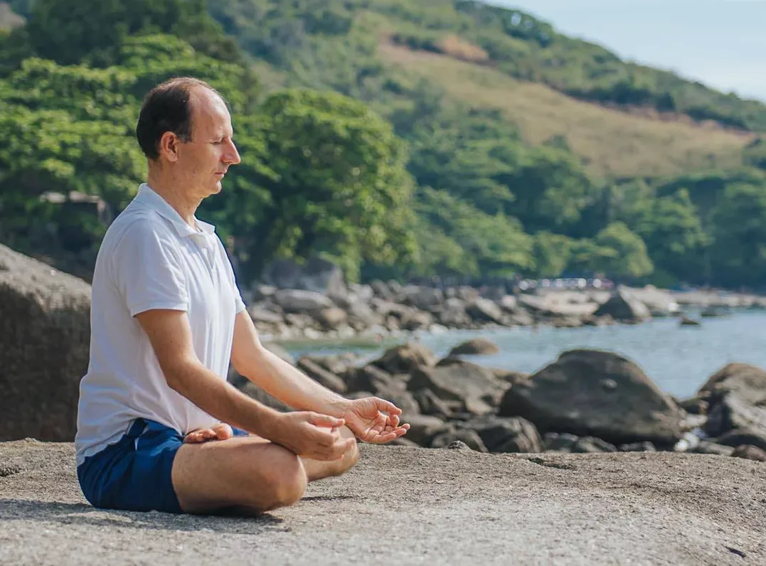 Remo Rittiner sitzt meditierend am Strand mit Felsen und Wasser im Hintergrund