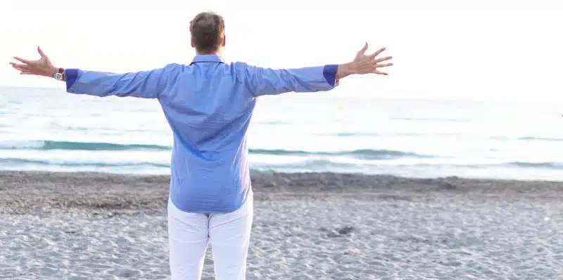 Mann in blauem Hemd mit ausgebreiteten Armen am Strand, Rückansicht