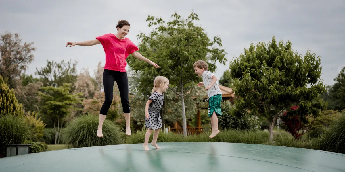 Frau springt auf Trampolin mit zwei Kindern im Garten, alle in Sportkleidung