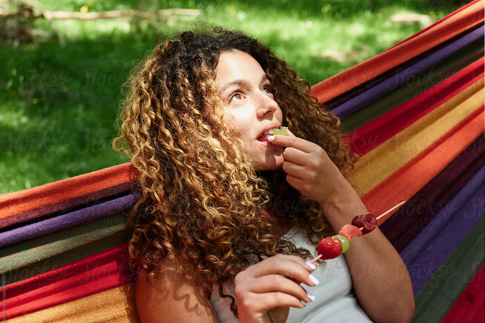Mädchen mit lockigem braunem Haar isst Beeren, sitzt auf farbigen Stoffen vor grünem Rasen