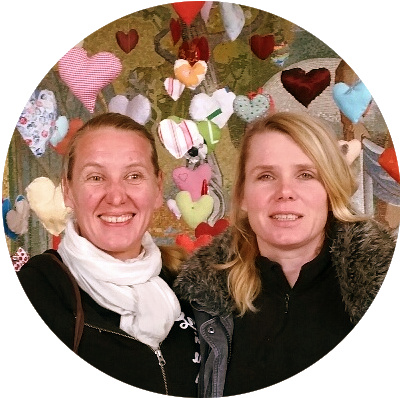 Porträtfoto von Katja Jessi, rundliches Format, zwei Frauen vor dekorativem Hintergrund mit Herzen und Blüten