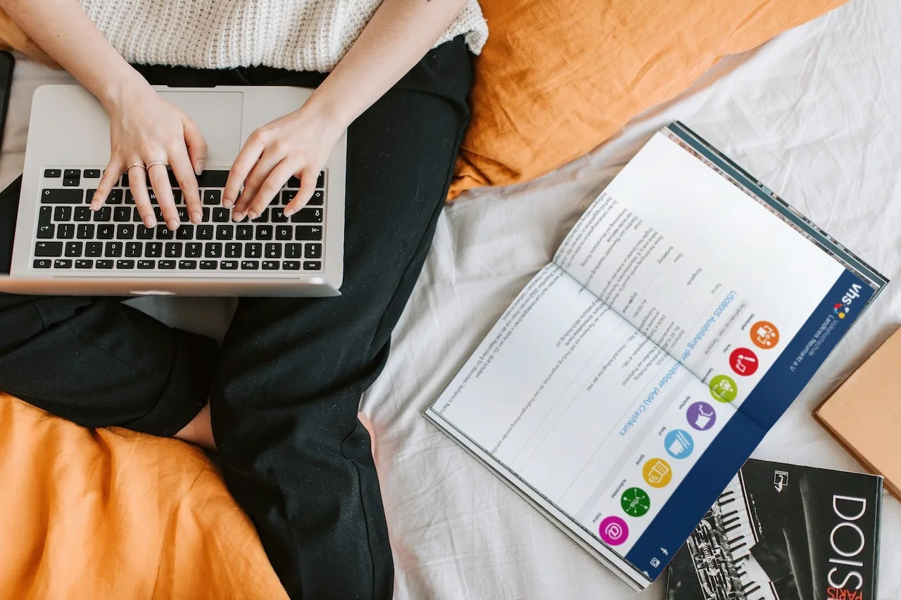Frauenhände auf Laptop-Tastatur, Person sitzt auf Couch, orangefarbene Decke sichtbar, Lernmaterial daneben, Online-Lernen