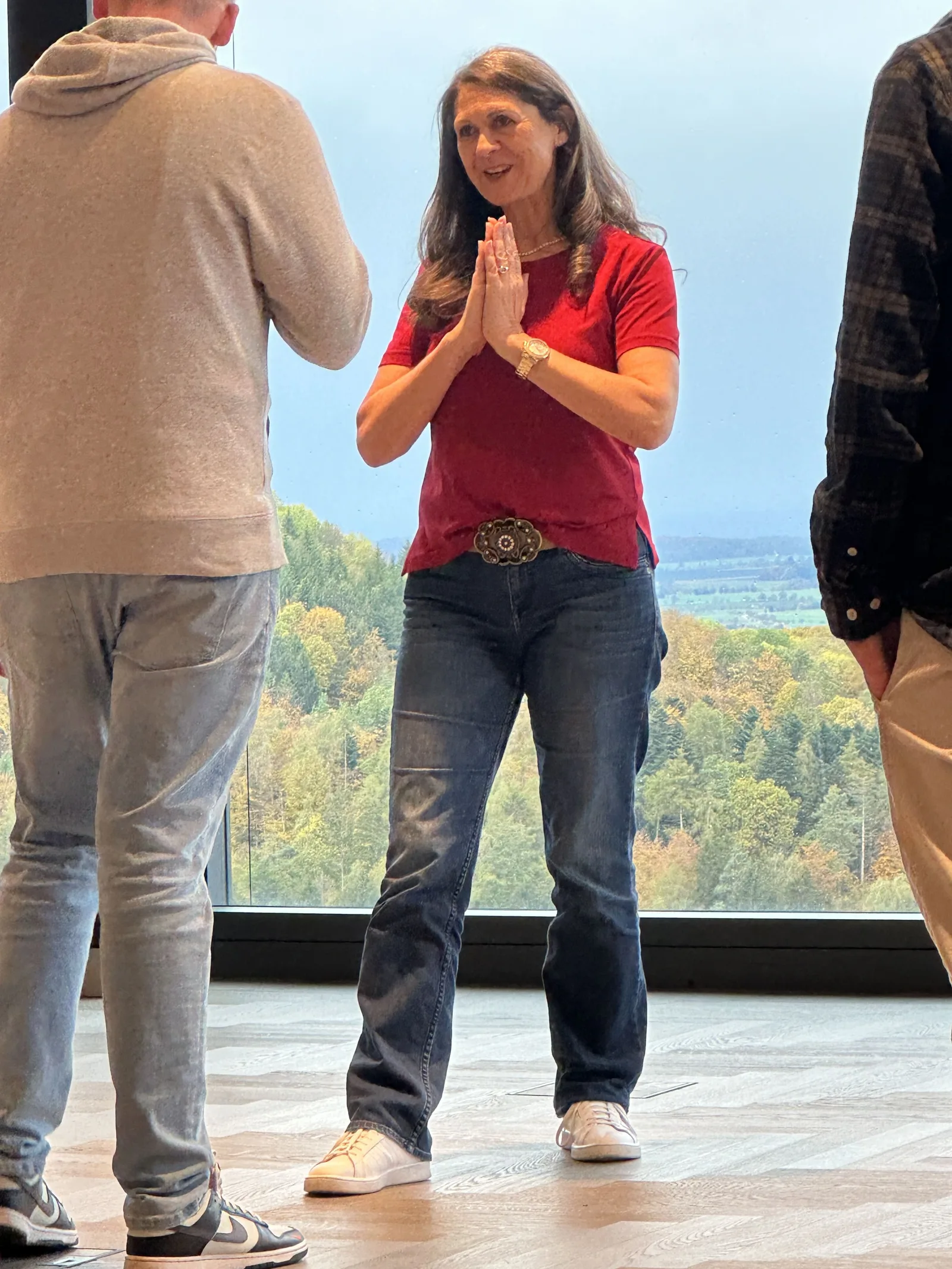 Frau in rotem Shirt und Jeans spricht zu Gruppe vor Panoramafenster mit Waldlandschaft, Praxis-Camp in kleiner Gruppe