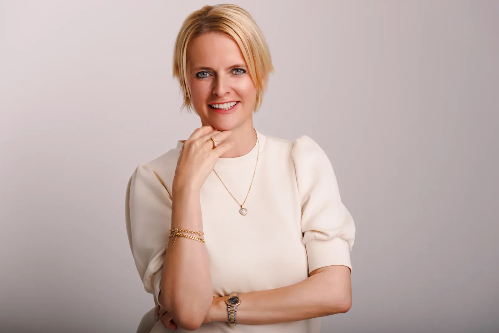 Gabriela Novák in cremefarbenem Pullover mit Gold-Schmuck, lächelndes Portrait vor beigem Hintergrund