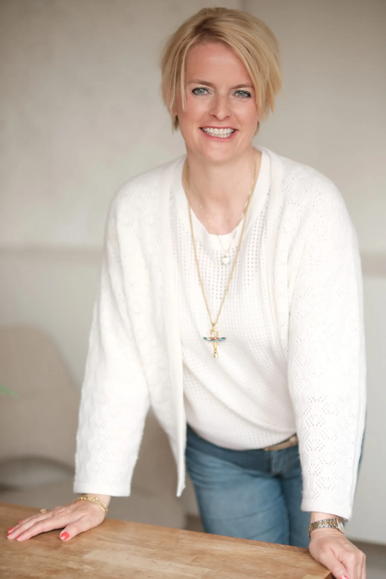 Gabriela Novák in weißer Strickjacke mit Kreuzamulett, lehnt an Holztisch, warmes Portrait
