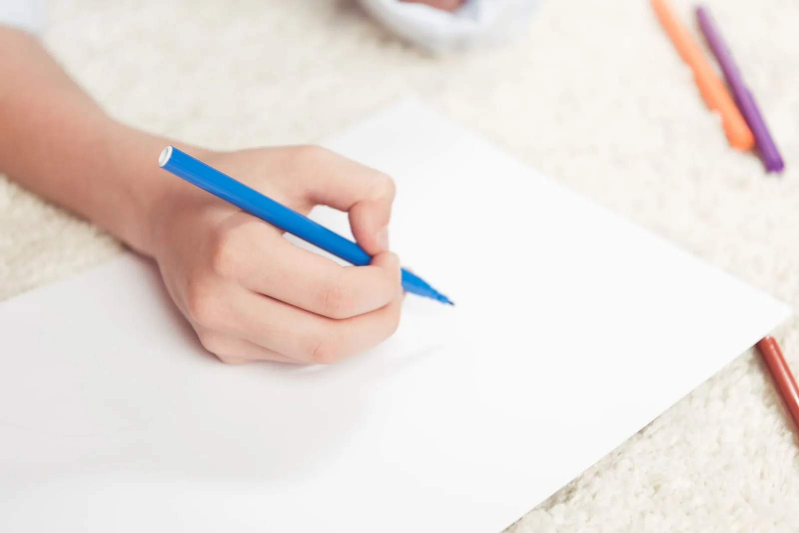 Kinderhand hält blauen Stift über Papier mit bunten Markern im Hintergrund
