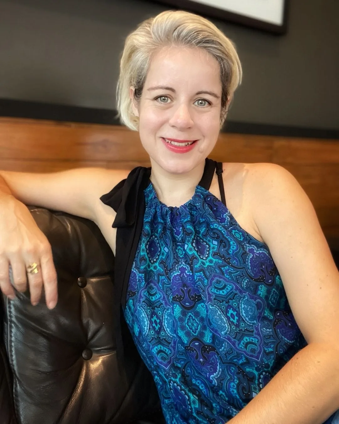 Nadja Teege lächelnd auf Sofa, blondes Haar, blaues Muster-Oberteil mit schwarzer Schärpe