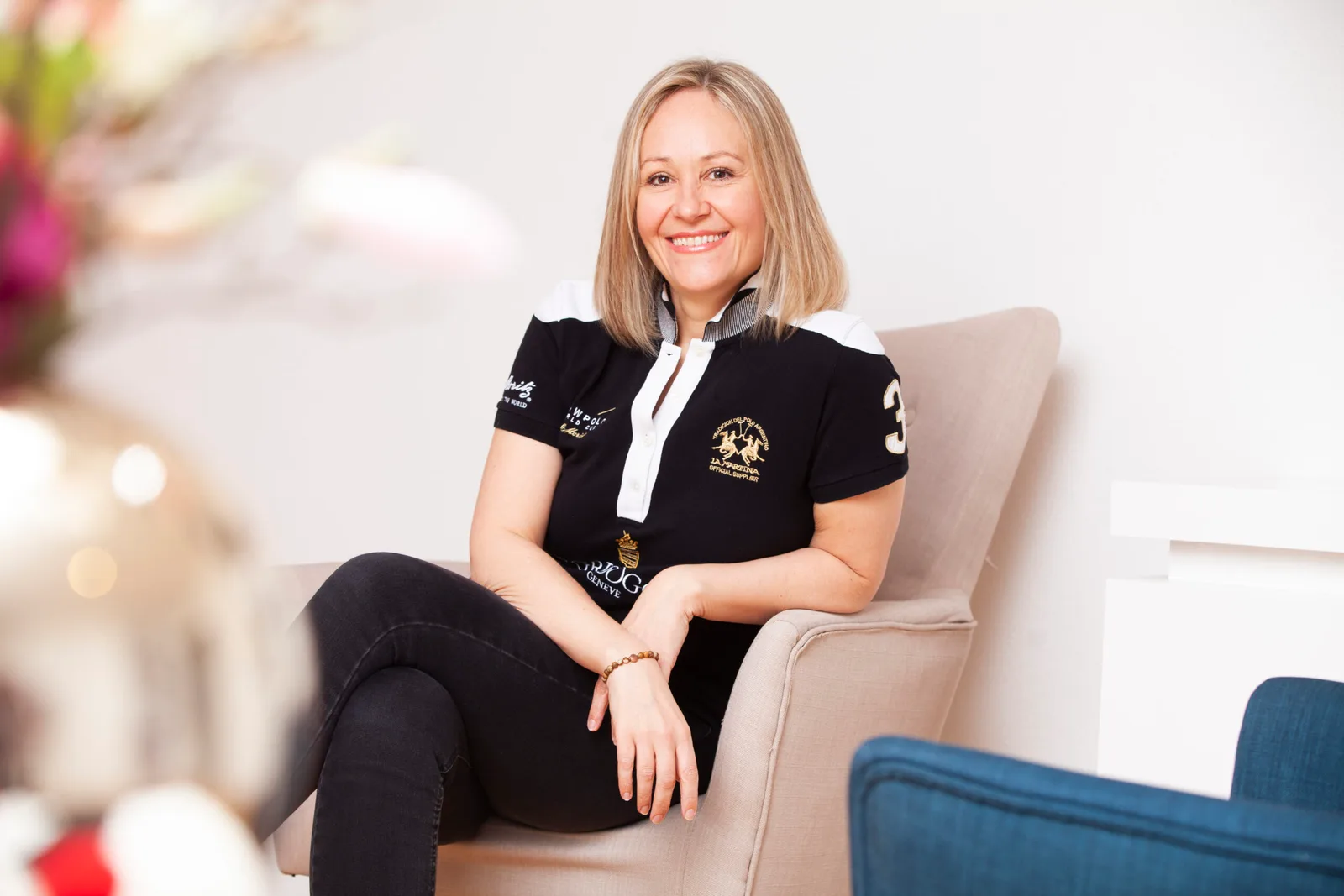 Anja Hussong sitzt auf hellem Sessel, schwarzes Poloshirt mit Logos, verschwommener Blütenhintergrund