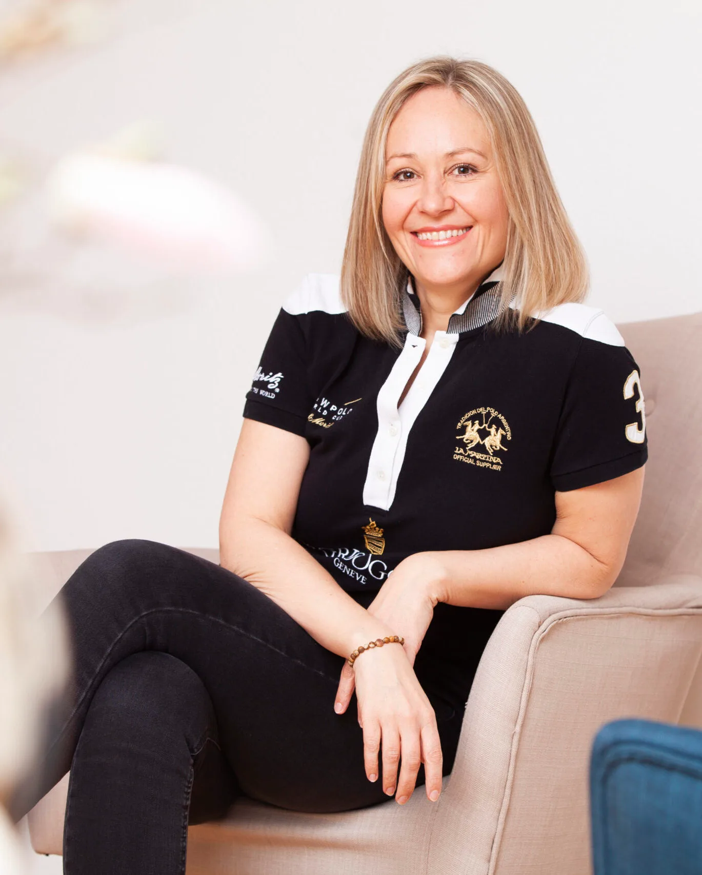 Anja Hussong sitzt lächelnd auf Sessel, trägt schwarzes Poloshirt mit Logo und weißem Kragen