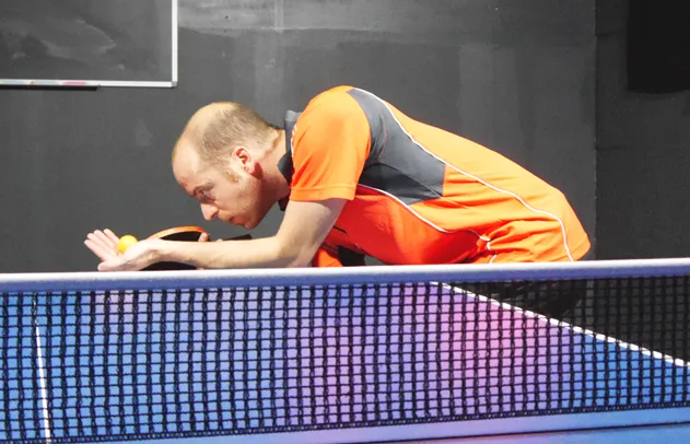 Tischtennisspieler Lars beim Aufschlag über der Tischtennisplatte in gelb-oranger Sportkleidung