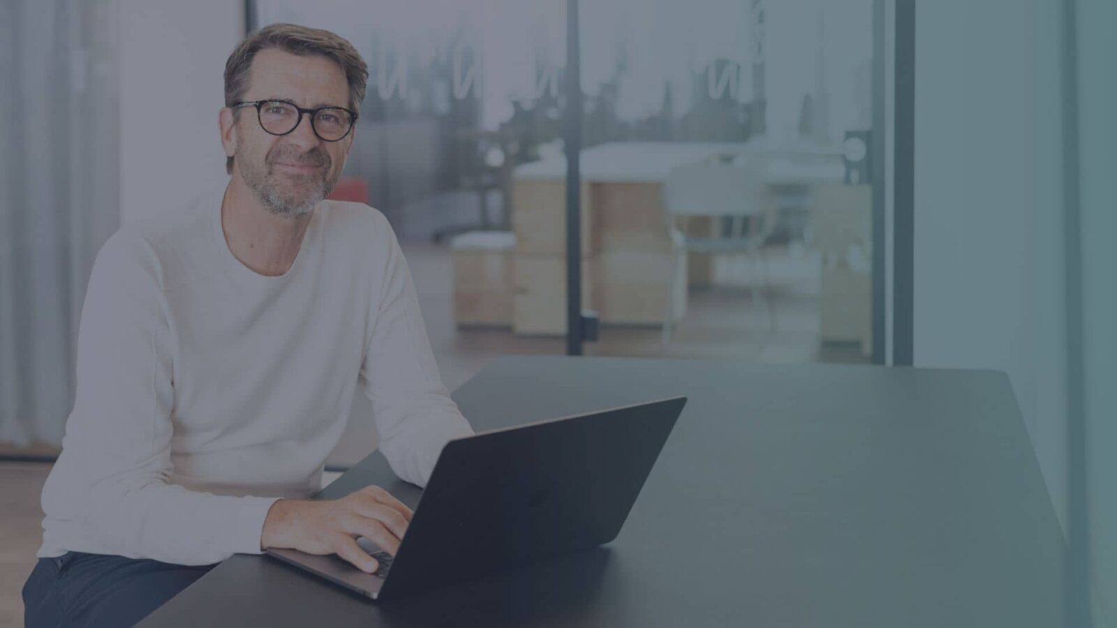 Mann mit Brille am Laptop in modernem Büro mit Fensterfront und Stadtblick