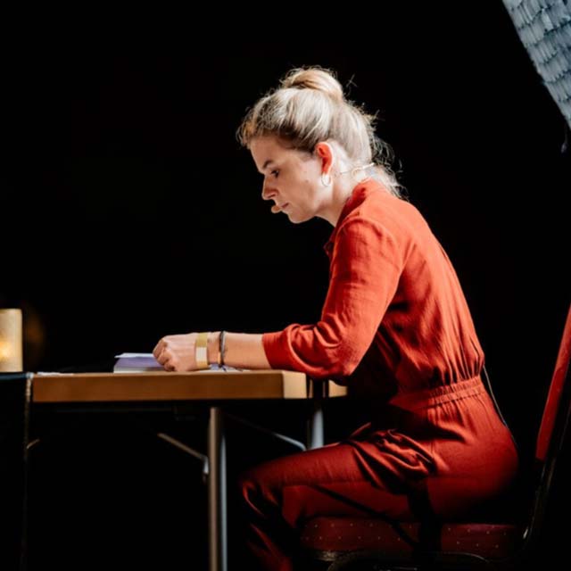 Frau in rotem Overall sitzt konzentriert an einem Tisch vor schwarzem Hintergrund