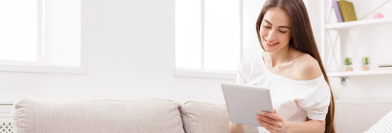Frau sitzt auf Sofa und schaut auf Tablet, helles Wohnzimmer mit Fenster und Regal