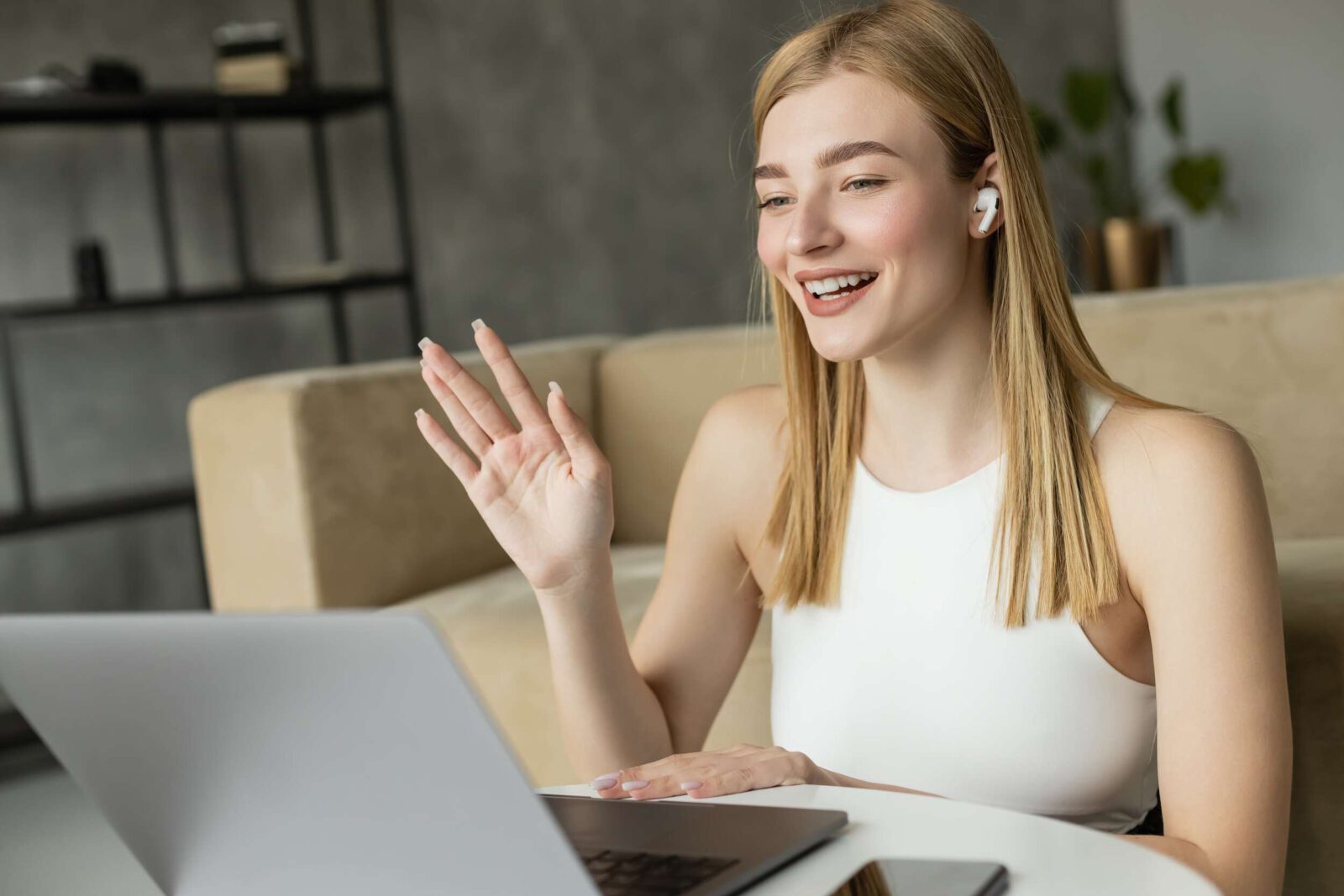 Junge Frau mit Kopfhörern winkt fröhlich in Laptop-Kamera, Online-Kommunikation