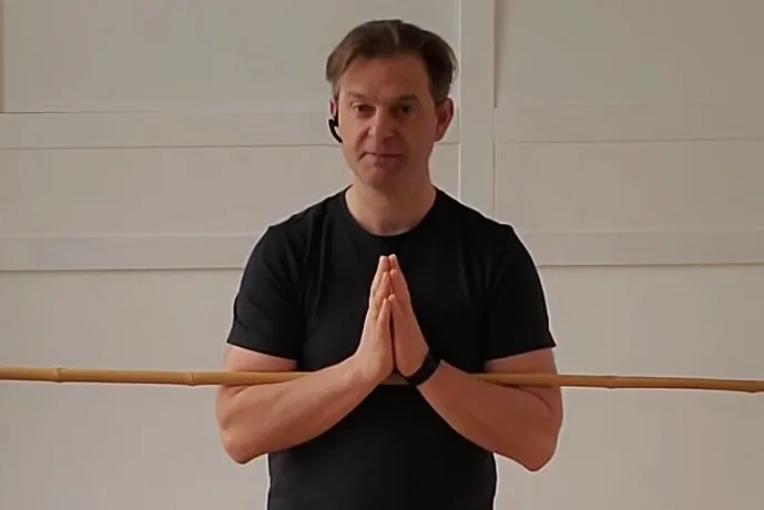 Mann in schwarzem Shirt mit Bambusstab vor weißer Wand in Grußposition