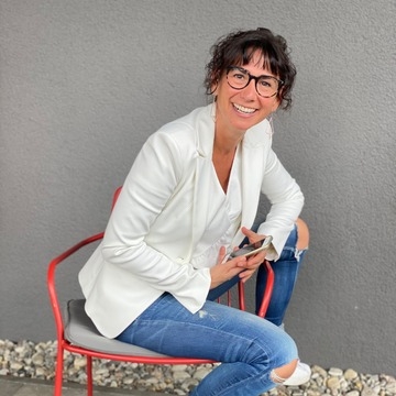 be ORIGINAL – Videokurs zur Positionierung von Monika Frauendorfer 12 Kundenstimme: Frau in weißer Bluse und Jeans sitzt lächelnd auf roten Stuhl vor grauem Hintergrund
