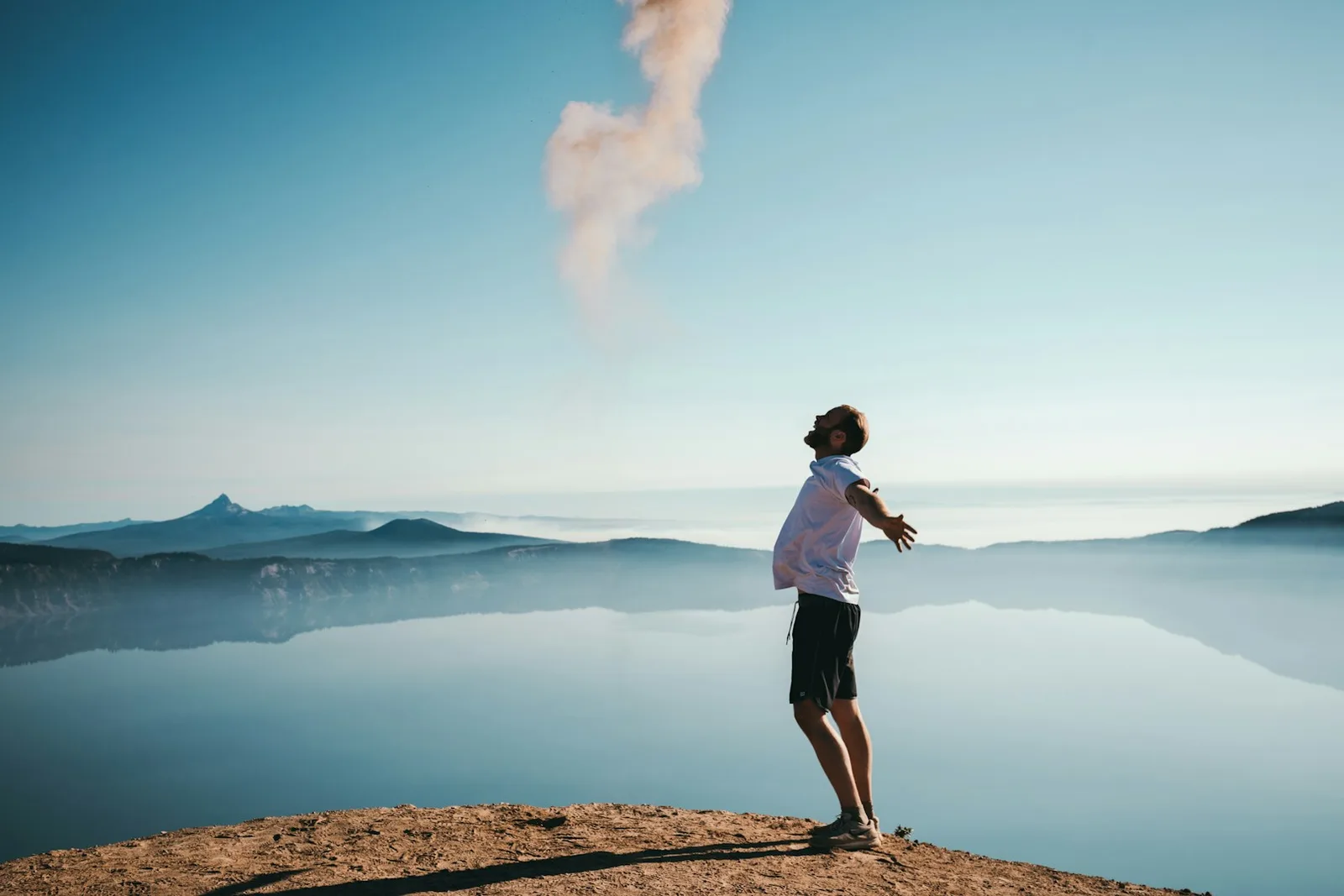 Mann in weißem Shirt und schwarzer Shorts steht auf Berghang mit Blick auf Bergsee und Berglandschaft