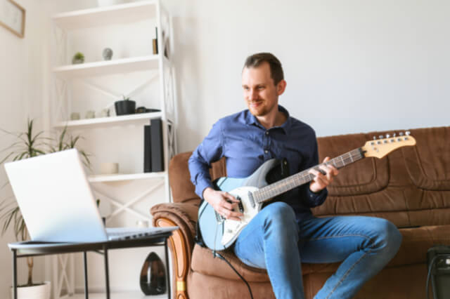Mann in blauem Hemd sitzt mit Gitarre auf Sofa vor Laptop
