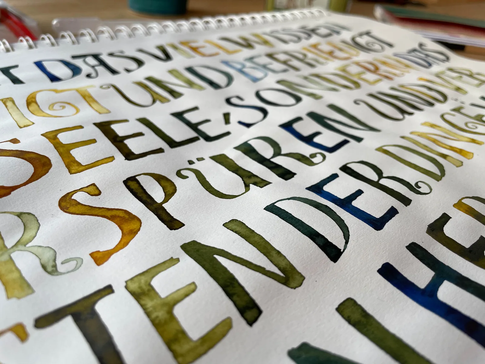 Handgeschriebene bunte Buchstaben auf Spiralblock, Kalligrafie-Übung mit Gold-, Grün- und Blautönen