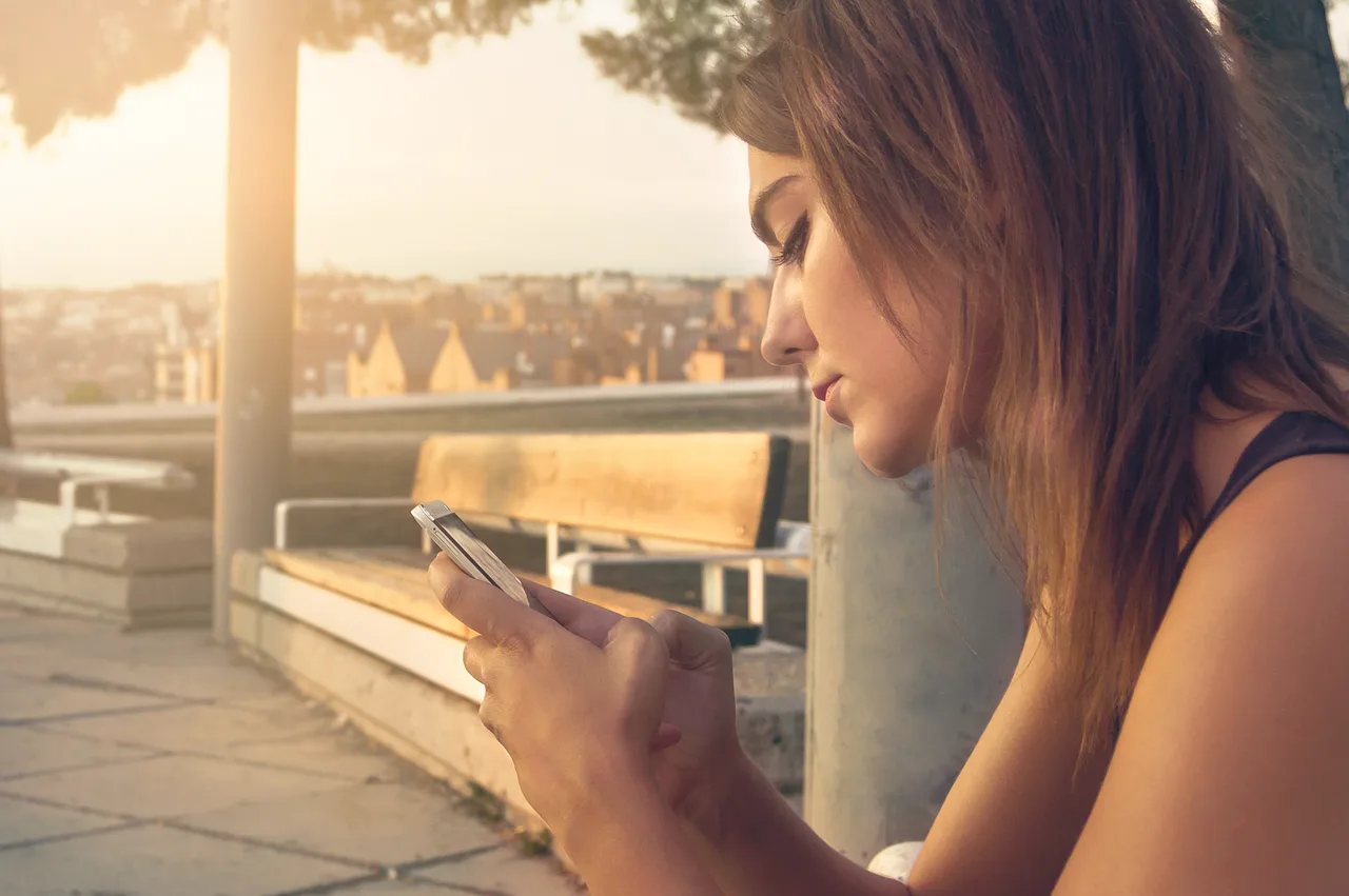 Frau auf Dachterrasse mit Smartphone bei Sonnenuntergang, urbane Skyline im Hintergrund