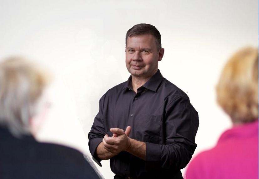 Uwe Krämer beim Coaching mit Paar im Hintergrund, gestikuliert erklärend vor hellem Hintergrund