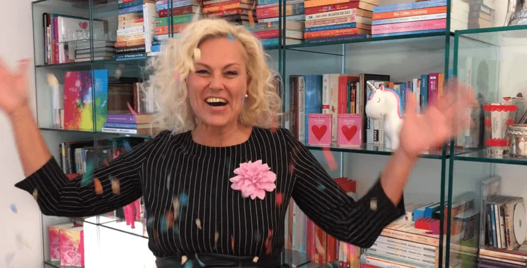 Blonde Frau in schwarzem Blazer mit rosa Blume vor Bücherregal, Arme oben ausgebreitet