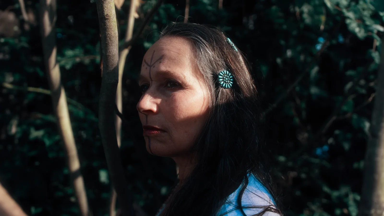 Naomi Pamuya Porträt von der Seite mit Teal Ohrrings im Wald vor dunklem Laub