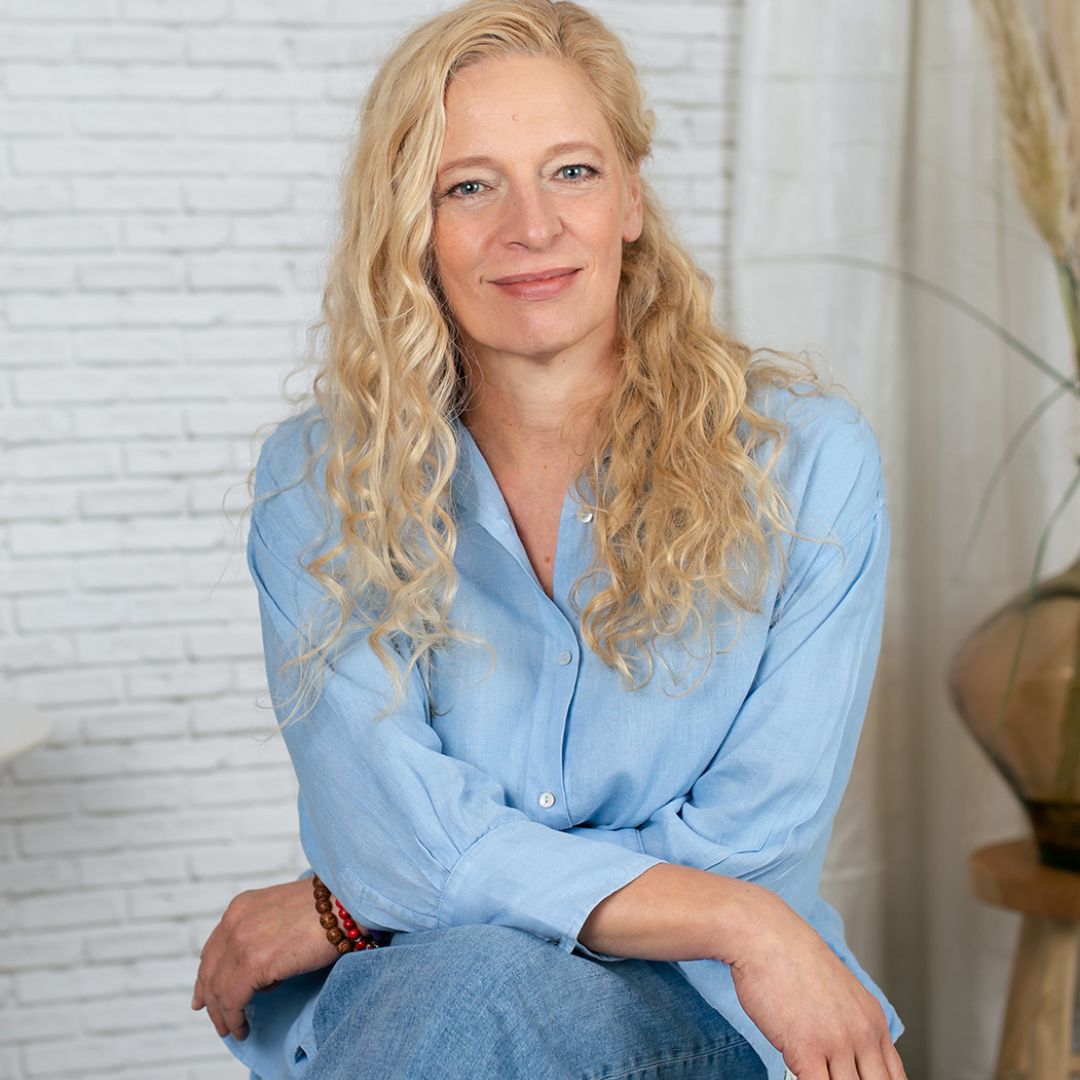Porträtfoto von Anja Engelsing, blonde Frau in hellblauem Hemd vor weißer Backsteinwand