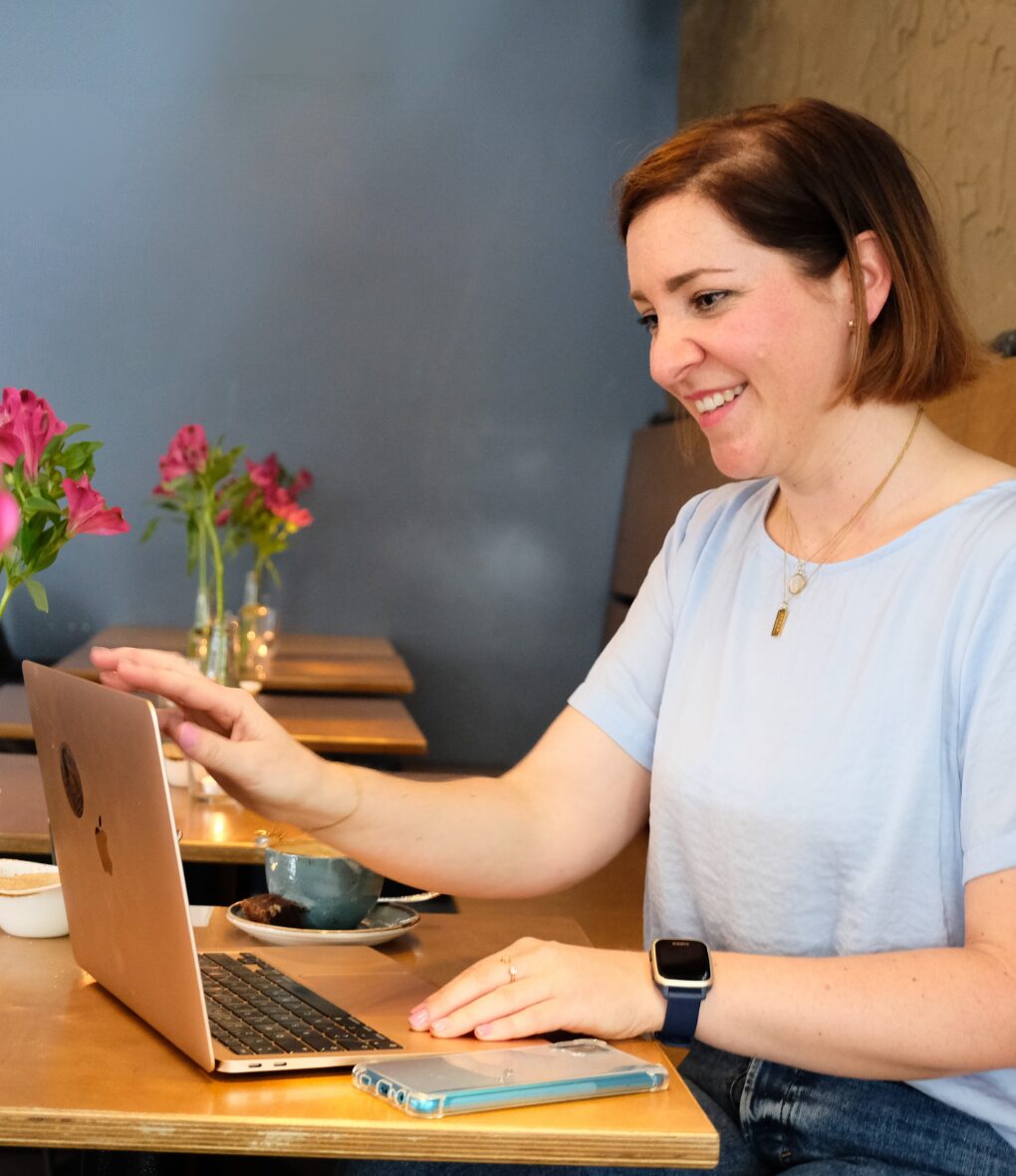 Frau mit rötlich-braunem Haar arbeitet lächelnd am Laptop, Blumen im Hintergrund
