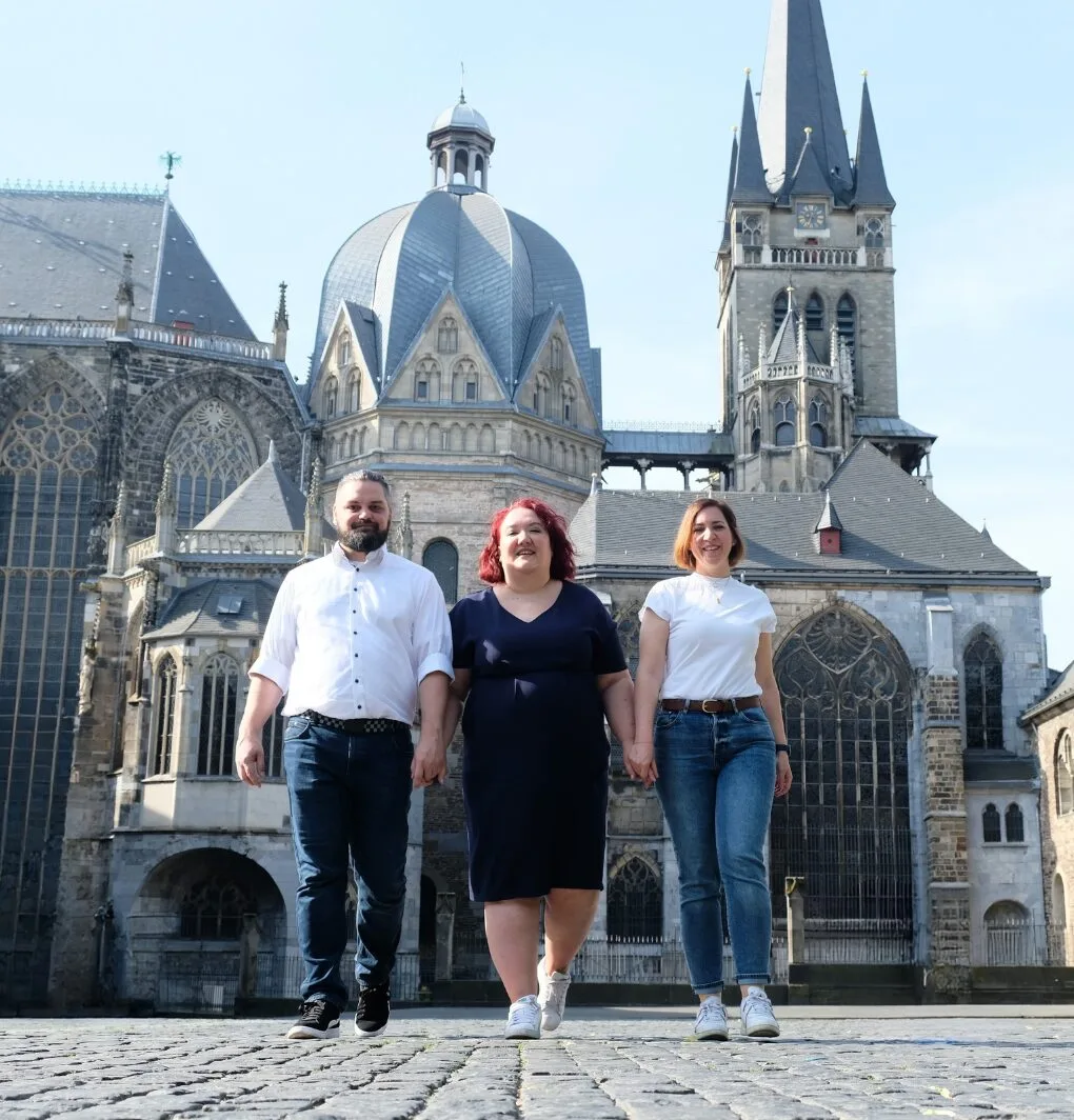 Drei Personen stehen vor einer gotischen Kirchenfassade, blauer Himmel