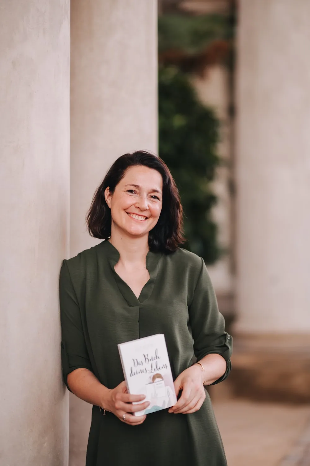 Buchmarketing Magie – Videokurs zur Autorenmarke 4 Sandy Mercier lächelnd in grünem Hemd mit Buch in den Händen vor heller Wand