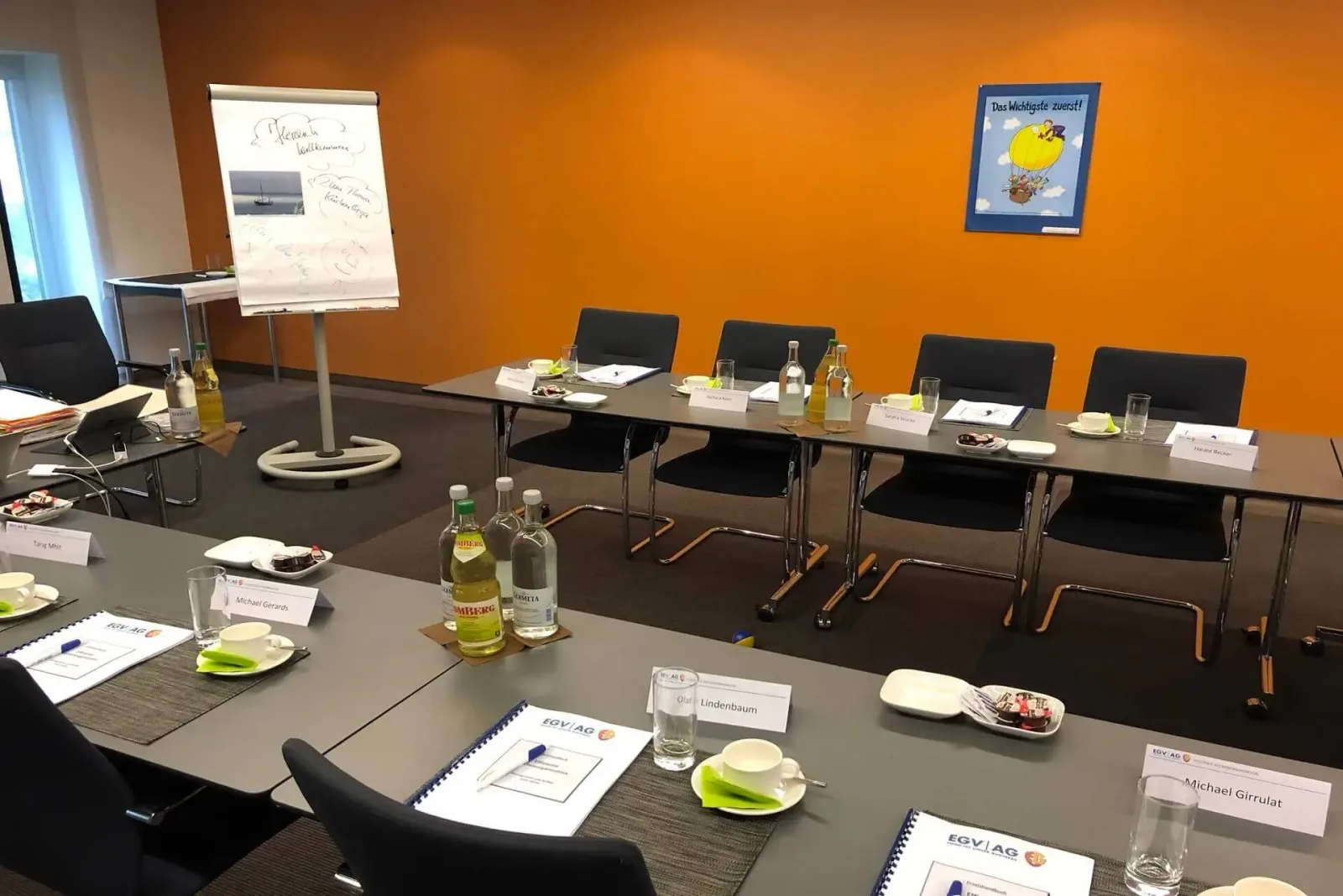 Seminarraum mit orangefarbener Wand, langer Tisch mit schwarzen Stühlen, Whiteboard und Getränken