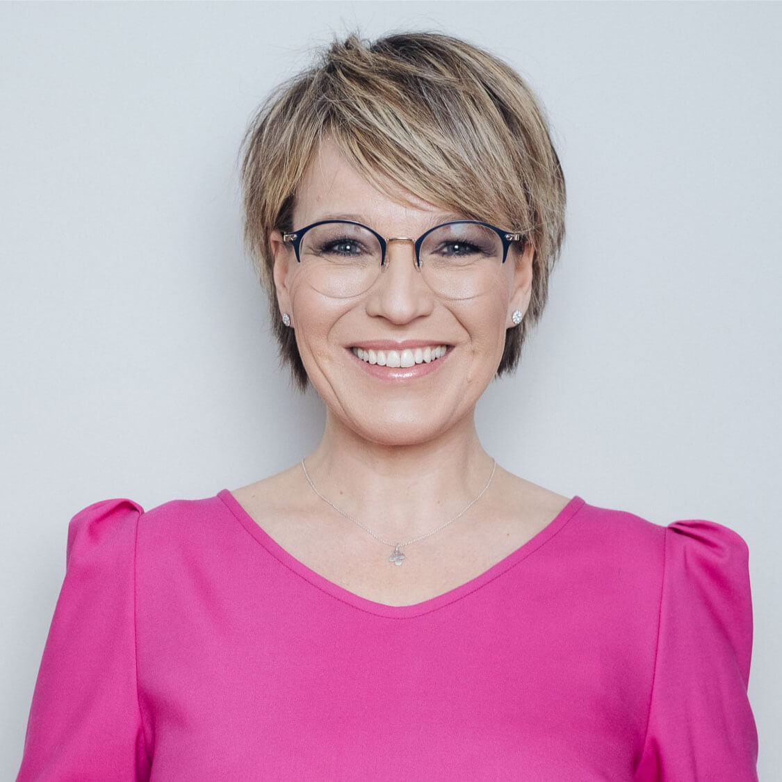 Porträt Frau mit Brille, kurzem blondem Haar, rosa Oberteil vor hellem Hintergrund, lächelnd