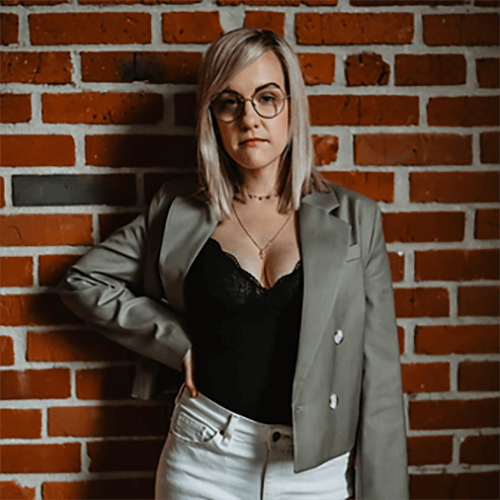 Porträt Frau mit Brille, grauem Blazer, schwarzem Top vor Backsteinhintergrund, selbstbewusste Pose