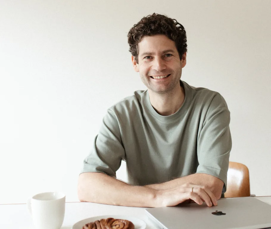Mann mit lockigem braunem Haar in hellgrünem T-Shirt sitzt lächelnd am weißen Tisch mit Kaffee und Gebäck
