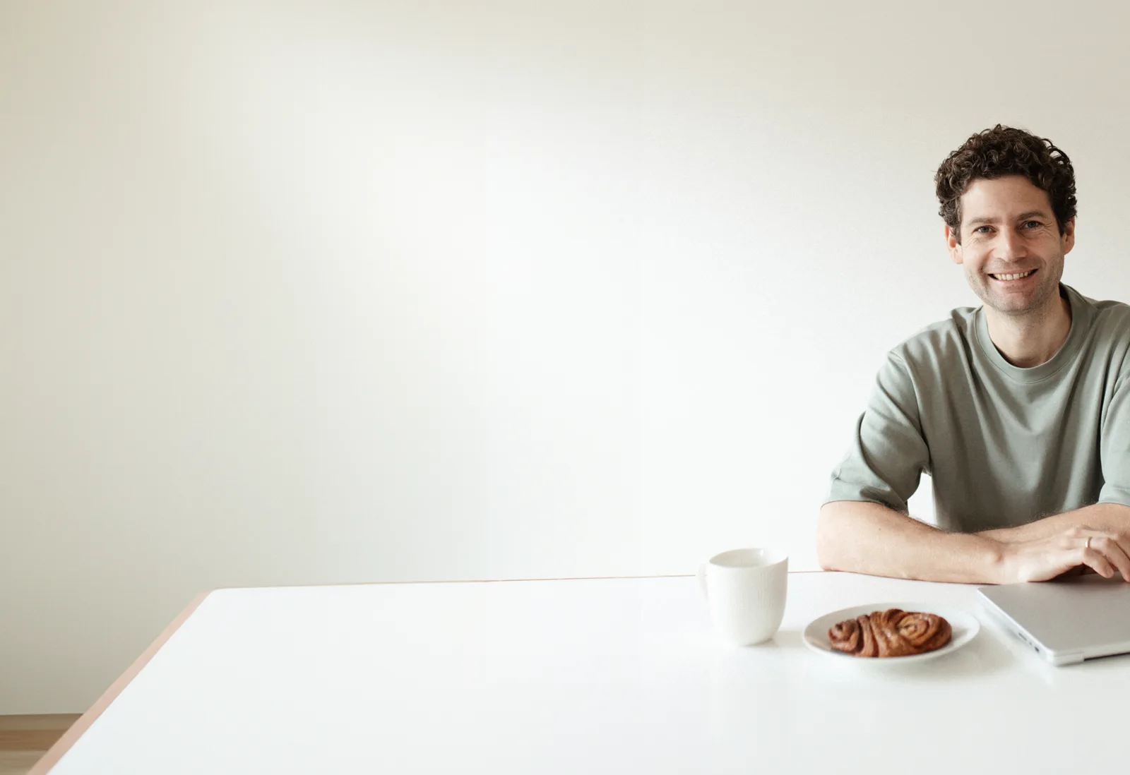 Mann am Tisch mit Kaffee und Keks, lächelt in die Kamera in minimalistischer Umgebung