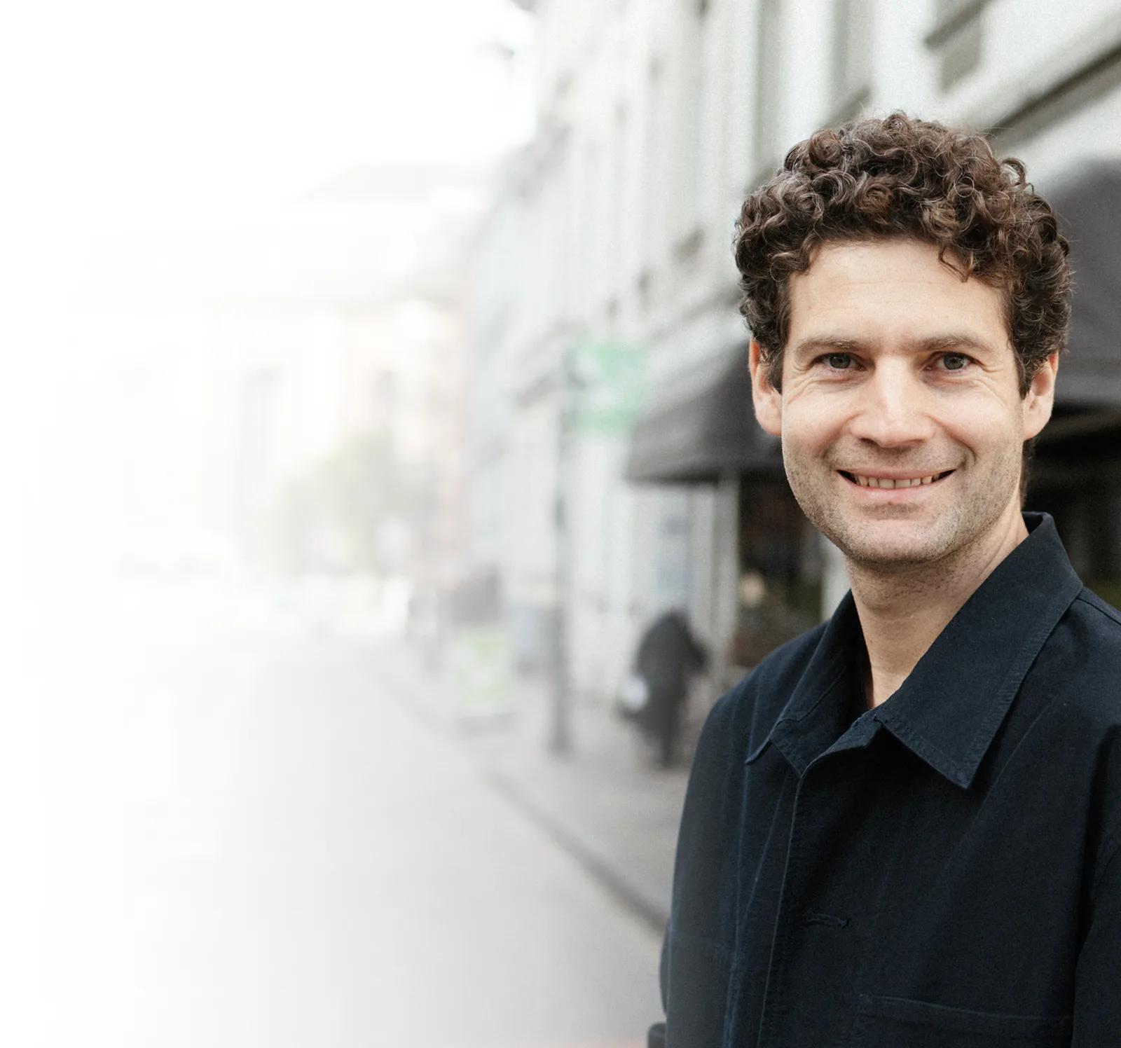 Portrait eines Mannes mit lockigen braunen Haaren in dunkler Jacke vor unscharfem Hintergrund
