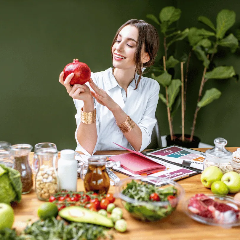 Ernährungsberaterin am Schreibtisch mit Obst und Gemüse, hält roten Apfel, grüner Hintergrund