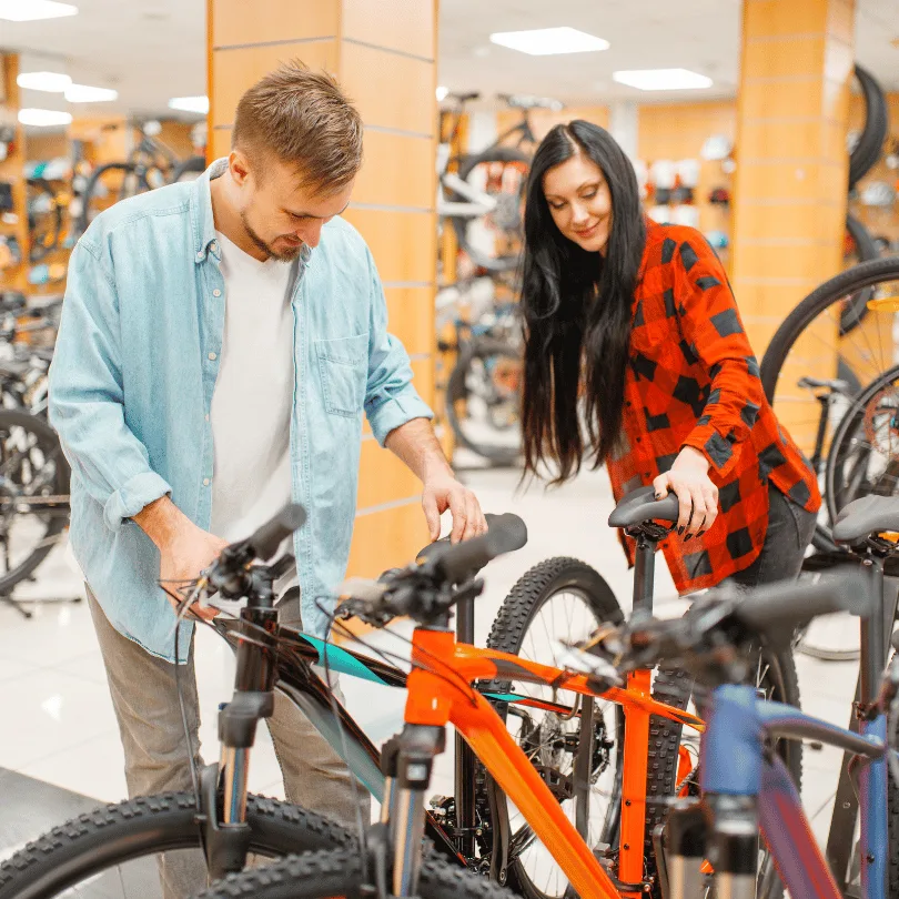 Mann und Frau in modernem Fahrradladen betrachten orangenes Mountainbike, helle Lagerbestände im Hintergrund