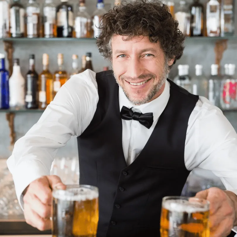 Businessplan Gastronomie/Kneipe – Vorlagenpaket mit Finanzplan 2 Bartender mit lockigem Haar und Fliege an der Bar, serviert zwei Bier-Gläser, Flaschenregal im Hintergrund