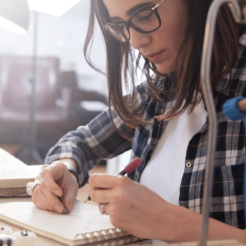 Businessplan Gold- und Silberschmied – Vorlage mit Finanzplan 2 Junge Frau mit Brille in kariertem Hemd arbeitet konzentriert an Schmuckstücken in der Werkstatt