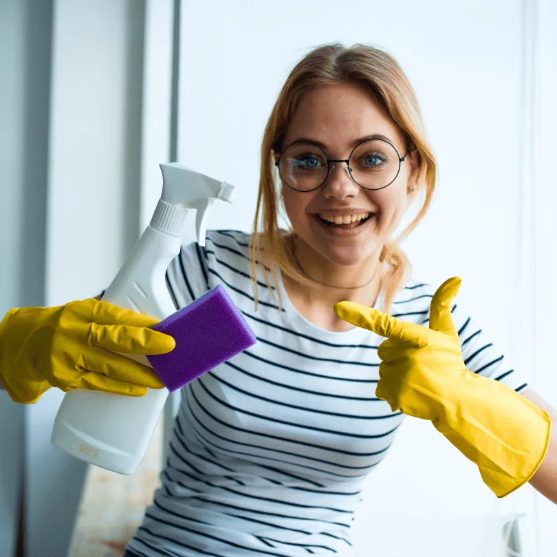 Businessplan Haushaltshilfe/Reinigungskraft von Dirk Leimkuhl 2 Junge Frau mit Brille, gelbem Putzbekleidung, Sprühflasche und Schwamm in den Händen auf hellem Hintergrund