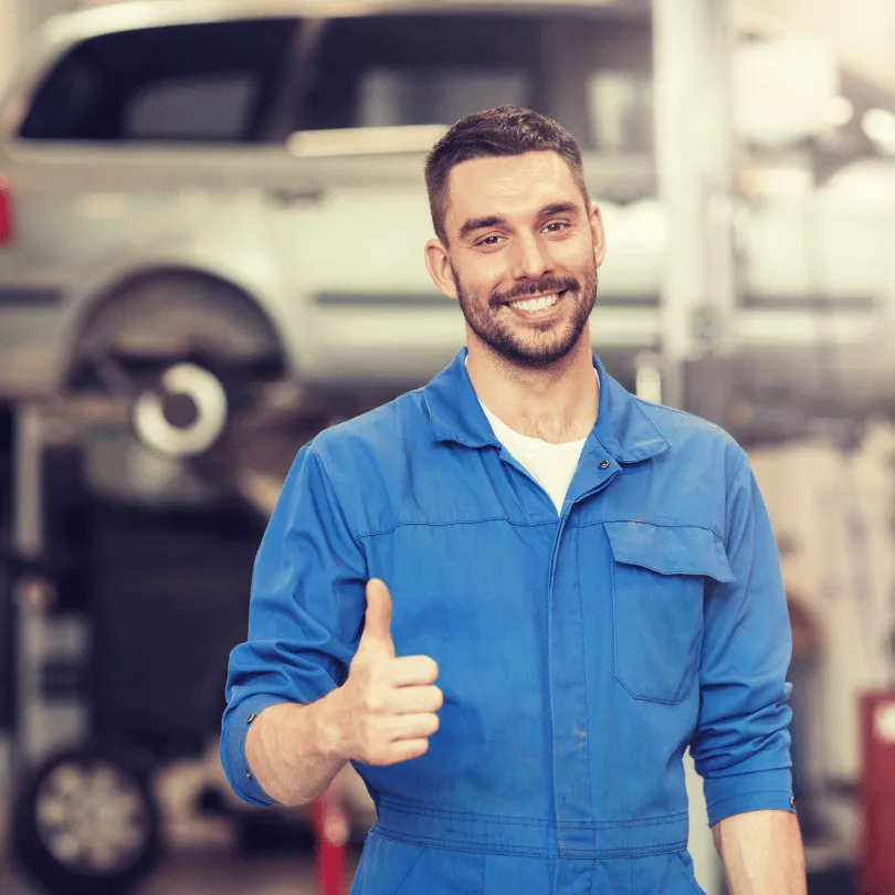 Junger Mechanic in blauer Werkstattkleidung mit Daumen-hoch-Geste in Autowerkstatt