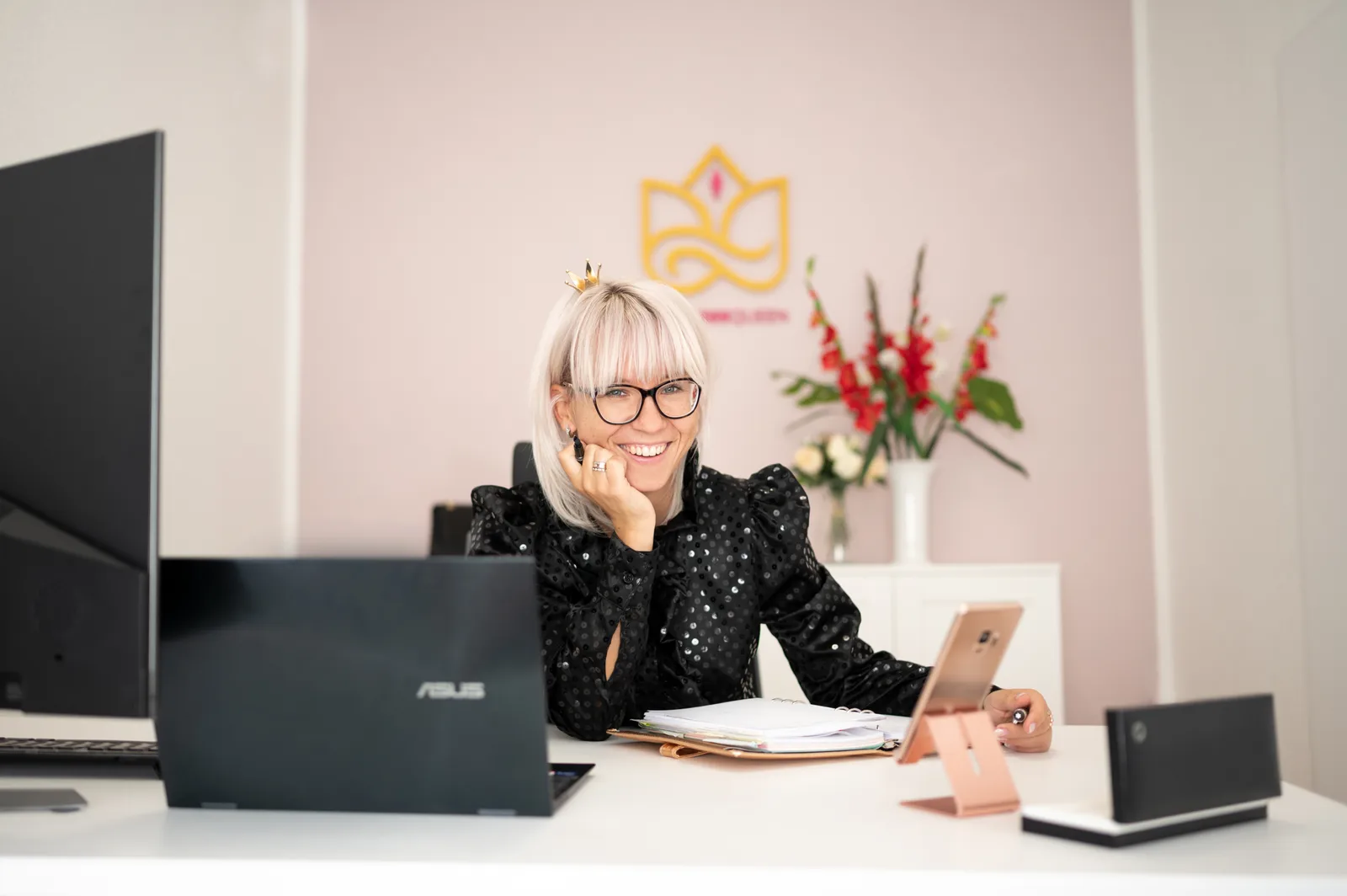 Julia Bock sitzt am Schreibtisch vor Laptop und Tablet, trägt schwarze Pailletten-Jacke, lächelt
