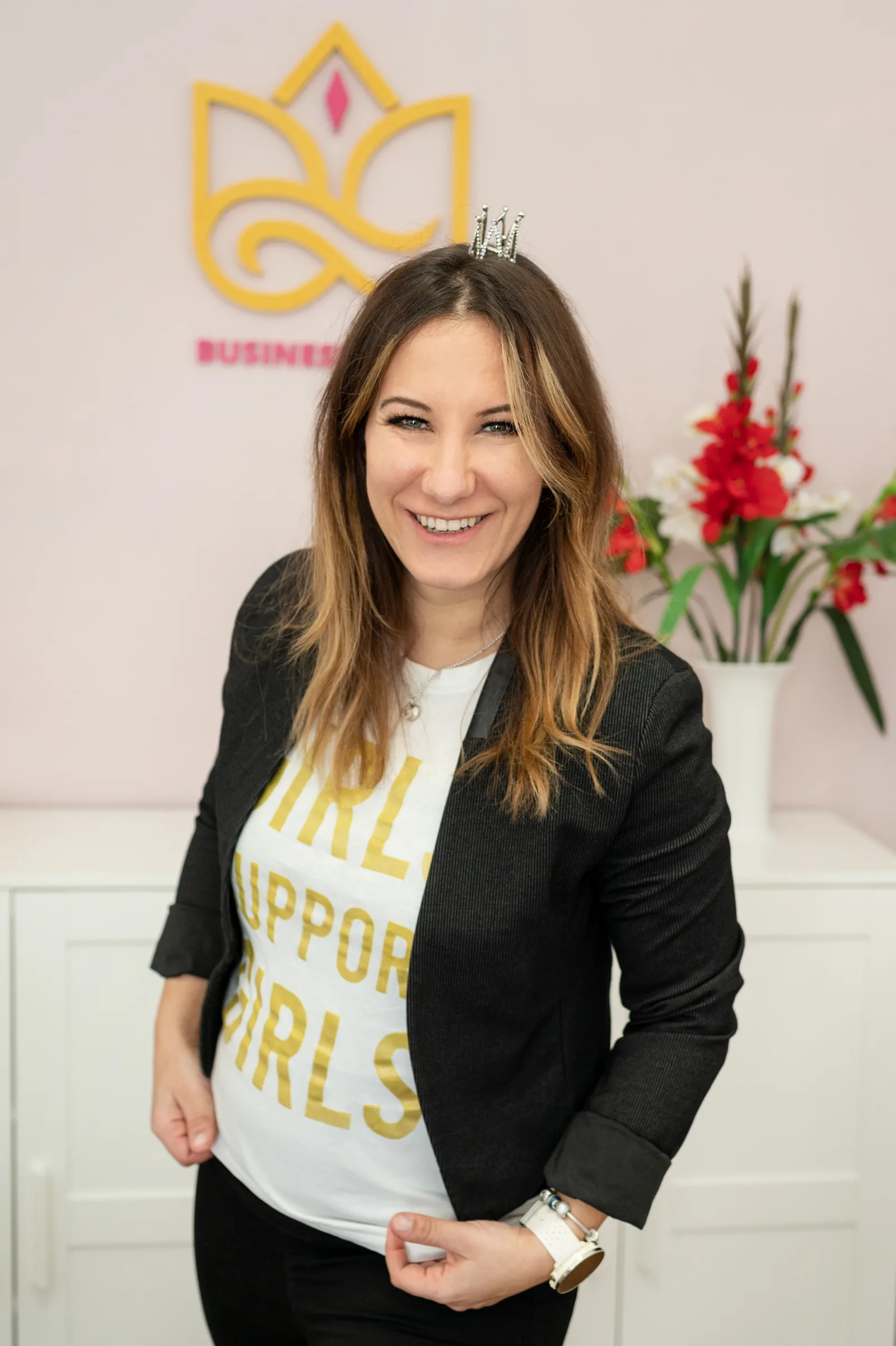 Frau mit braunem Haar, Tiara und Blazer vor gelbem Logo, trägt Shirt mit Goldtext
