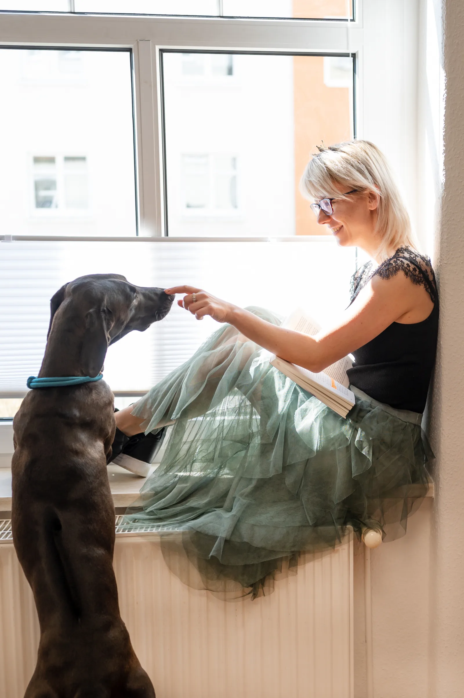 Frau mit weißblondem Haar und Brille sitzt am Fenster mit schwarzem Hund auf dem Schoß