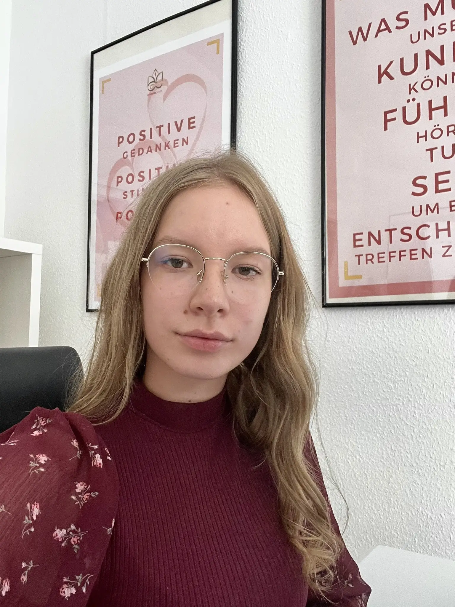 Junge Frau mit braunem Haar und Brille vor Motivationsplakaten, trägt burgunderroten Pullover