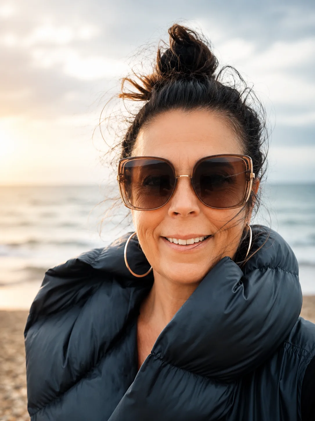 Porträt einer Frau mit Sonnenbrille und Dutt am Strand, lächelnd