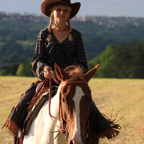 Anna Köhler in Cowboyhut sitzt auf braunem Pferd, Landschaft
