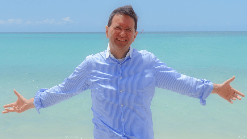 André Loibl am Strand mit blauem Hemd, Arme ausgebreitet, Meer im Hintergrund